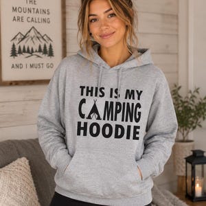 May include: A light gray hoodie with the text "THIS IS MY CAMPING HOODIE" in black. The hoodie has a front pocket and a drawstring hood. A framed sign with the text "THE MOUNTAINS ARE CALLING AND I MUST GO" is in the background.