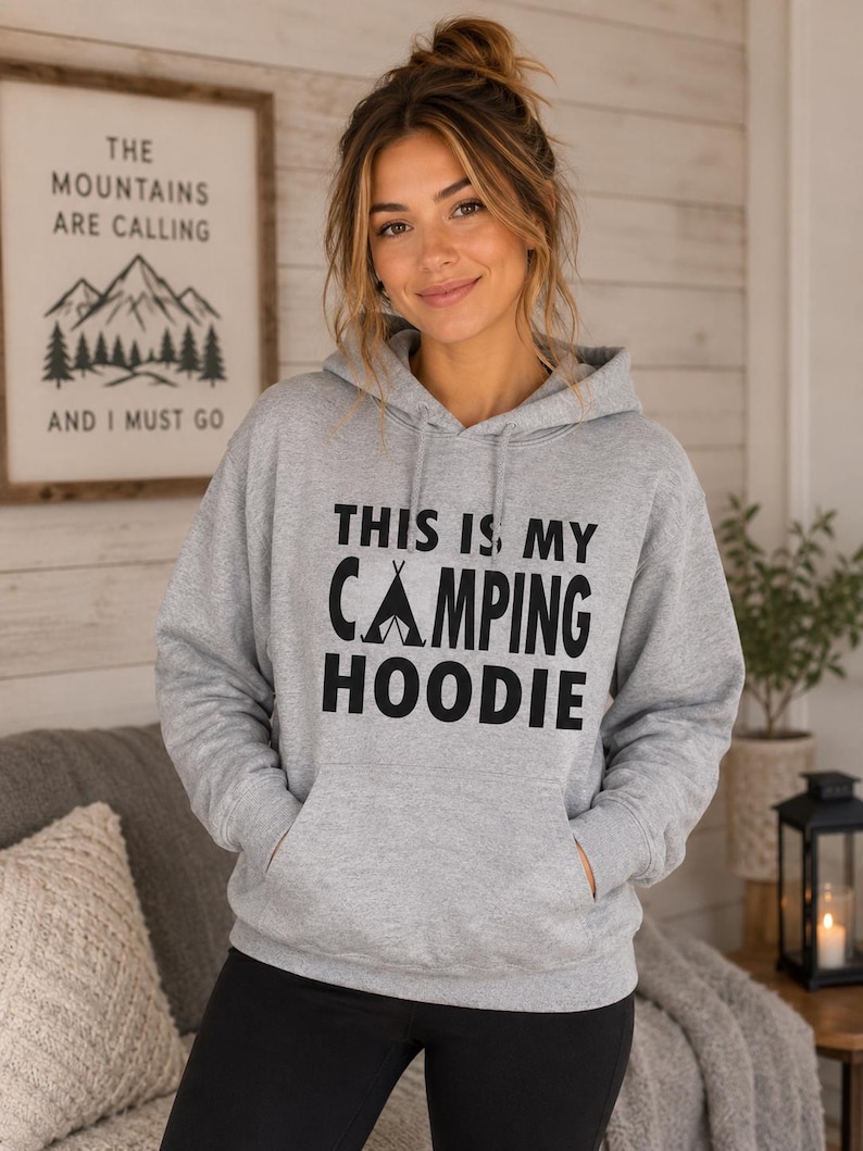 May include: A light gray hoodie with the text "THIS IS MY CAMPING HOODIE" in black. The hoodie has a front pocket and a drawstring hood. A framed sign with the text "THE MOUNTAINS ARE CALLING AND I MUST GO" is in the background.