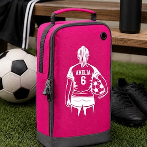 May include: A vibrant pink soccer boot bag with grey trim, displaying a white graphic of a soccer player with the name "AMELIA" and the number "6". A black water bottle, football, and boots are in the background.