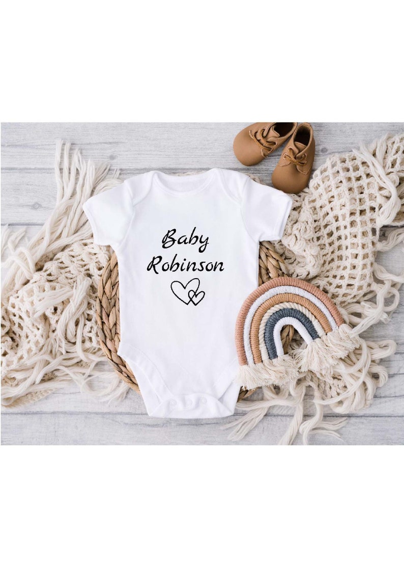 Może przedstawiać: Biały śpioszek dla niemowlęcia z czarnym napisem "Baby Robinson" i dwoma czarnymi sercami.