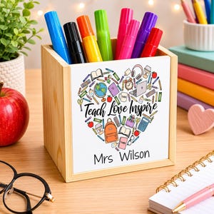 May include: A wooden desk organizer with a white front panel featuring a heart-shaped design filled with school-related illustrations. The text "Teach Love Inspire" and "Mrs Wilson" are printed on the panel. The organizer holds colorful markers.