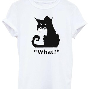 May include: White t-shirt featuring a black silhouette of an angry cat with the text "What?". The cat has pointed ears, a curled tail, and a grumpy expression. The shirt is hanging on a wooden hanger.