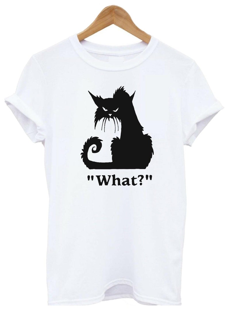 May include: White t-shirt featuring a black silhouette of an angry cat with the text "What?". The cat has pointed ears, a curled tail, and a grumpy expression. The shirt is hanging on a wooden hanger.