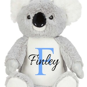 Puede incluir: Un peluche de koala con suave pelaje gris, orejas interiores blancas y almohadillas blancas en los pies. El koala tiene una nariz negra grande y ojos negros. Un parche blanco en el pecho muestra una letra F azul claro y el nombre Finley en negro.