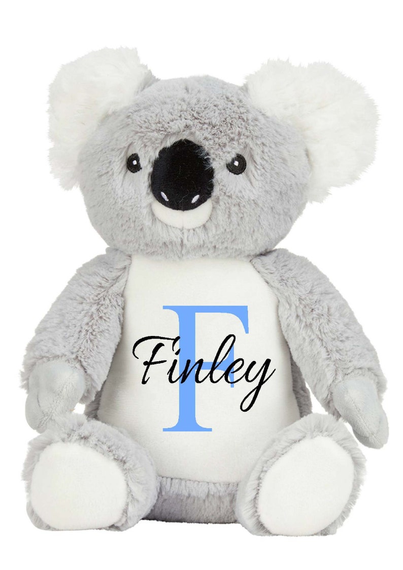 Puede incluir: Un peluche de koala con suave pelaje gris, orejas interiores blancas y almohadillas blancas en los pies. El koala tiene una nariz negra grande y ojos negros. Un parche blanco en el pecho muestra una letra F azul claro y el nombre Finley en negro.