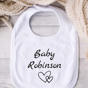 Może przedstawiać: Biały śliniaczek dla niemowlęcia z czarnym napisem "Baby Robinson" i czarnym wzorem serca.