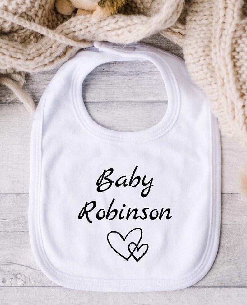 Może przedstawiać: Biały śliniaczek dla niemowlęcia z czarnym napisem "Baby Robinson" i czarnym wzorem serca.