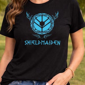 May include: Black t-shirt with a blue graphic design. The design includes a shield-like emblem and the word "SHIELDMAIDEN" in a stylized font. The design is centered on the chest.