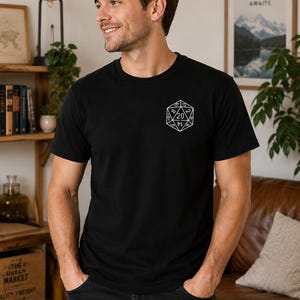 May include: A black short-sleeved t-shirt featuring a white line art graphic of a twenty-sided die on the left chest. The die design displays various numbers, with the number 20 prominently featured at the top centre of the geometric shape.