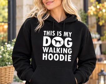 May include: A black hooded sweatshirt featuring white text that reads THIS IS MY DOG WALKING HOODIE with a silhouette of a dog replacing the letter O in the word DOG. The garment includes a front kangaroo pocket, long sleeves, and a drawstring hood.
