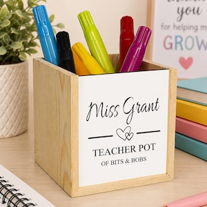May include: A square wooden pencil holder featuring a white front panel with the black printed text Miss Grant, a heart icon, and the words TEACHER POT OF BITS & BOBS. The container holds several colourful markers in blue, black, yellow, green, red, and purple.