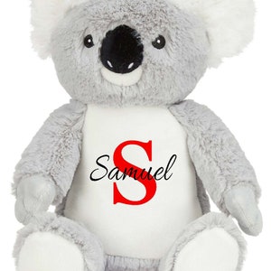 Puede incluir: Un peluche de koala con pelaje gris suave, mechones blancos en las orejas y nariz negra. El koala presenta un parche blanco en el vientre impreso con una letra S roja grande y el nombre Samuel en letra cursiva negra en el centro.