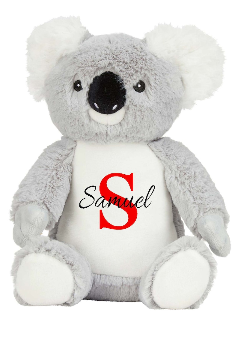 Puede incluir: Un peluche de koala con pelaje gris suave, mechones blancos en las orejas y nariz negra. El koala presenta un parche blanco en el vientre impreso con una letra S roja grande y el nombre Samuel en letra cursiva negra en el centro.
