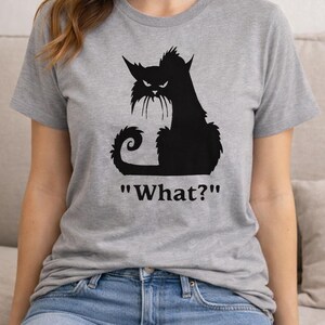 May include: A gray t-shirt featuring a black silhouette of an angry cat with the text "What?" below. The cat has pointed ears, a curled tail, and a grumpy expression. The shirt is made of a soft material.