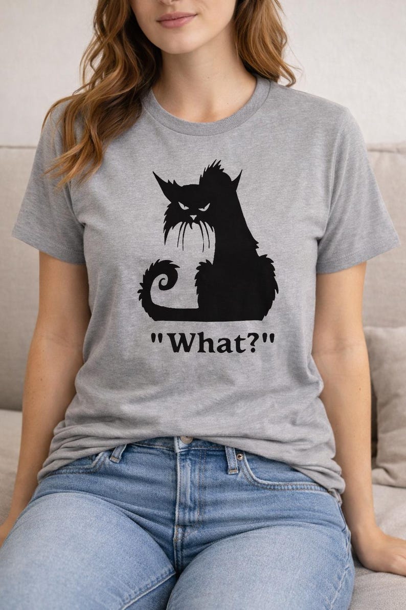 May include: A gray t-shirt featuring a black silhouette of an angry cat with the text "What?" below. The cat has pointed ears, a curled tail, and a grumpy expression. The shirt is made of a soft material.