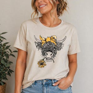 May include: Beige t-shirt featuring a black and white illustration of a Highland cow with a yellow polka-dot bandana and a sunflower in its mouth. The cow has long, shaggy fur and curved horns. The shirt is made of a soft material.