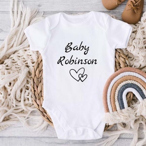 Może przedstawiać: Biały śpioszek dla niemowlęcia z napisem "Baby Robinson" i dwoma splecionymi sercami.