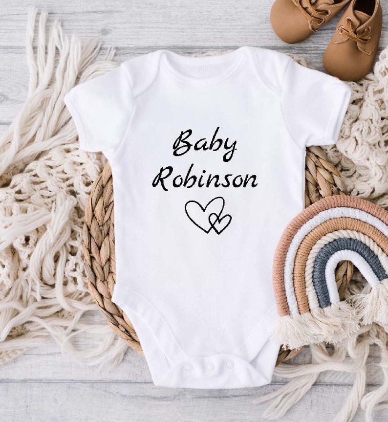 Może przedstawiać: Biały śpioszek dla niemowlęcia z napisem "Baby Robinson" i dwoma splecionymi sercami.
