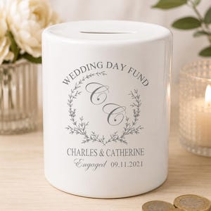Könnte beinhalten: Weiße Keramik-Sparbüchse für den Hochzeitstag mit einem Schlitz oben. Die Sparbüchse hat ein graues Kranzdesign mit den Initialen "C" und dem Text "Wedding Day Fund" und "Charles & Catherine Engaged 09.11.2021".