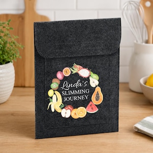 May include: A dark gray felt pouch with a flap closure. The pouch features a colorful fruit and vegetable wreath design with the text "Linda's Slimming Journey". The design includes bananas, oranges, and watermelon.