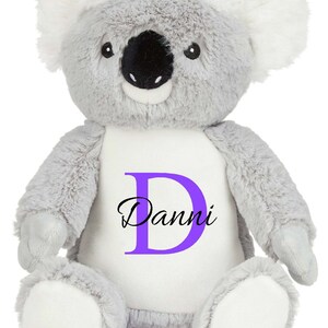 Puede incluir: Un peluche de koala con pelaje gris suave, orejas interiores blancas y una mancha blanca en el vientre. El vientre presenta una letra D grande de color morado con el nombre Danni impreso en negro en letra cursiva. Tiene nariz negra.