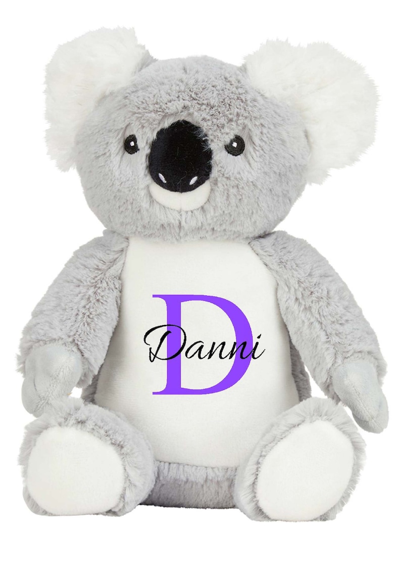 Puede incluir: Un peluche de koala con pelaje gris suave, orejas interiores blancas y una mancha blanca en el vientre. El vientre presenta una letra D grande de color morado con el nombre Danni impreso en negro en letra cursiva. Tiene nariz negra.