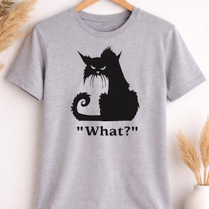 May include: A light gray t-shirt featuring a black silhouette of a cat with an angry expression. The cat has pointed ears and a curled tail. Below the cat is the text "What?" in black.