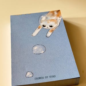 May include: A blue notepad with a cartoon cat illustration. The cat is orange, white, and brown. The notepad has the text "COSMOS OF OURS" at the bottom. The notepad is rectangular and has a stack of pages.