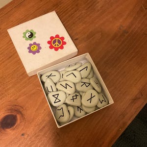 May include: A square white box containing a collection of smooth, light-colored stones marked with black runic symbols. The box lid is detached and features three colorful flower-shaped stickers: a yin-yang, a smiley face, and a peace sign.