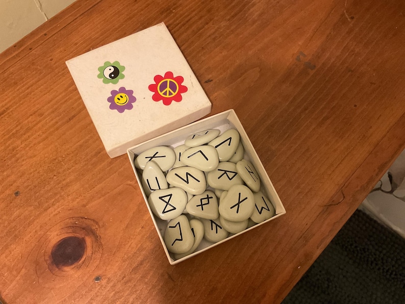 May include: A square white box containing a collection of smooth, light-colored stones marked with black runic symbols. The box lid is detached and features three colorful flower-shaped stickers: a yin-yang, a smiley face, and a peace sign.