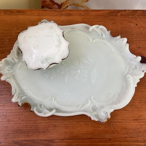 May include: A white porcelain tray with a decorative floral design and a raised edge. The tray has a scalloped edge and is in the shape of a heart. A small white porcelain dish sits on top of the tray.