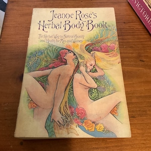 May include: A paperback book titled Jeanne Rose's Herbal Body Book: The Herbal Way to Natural Beauty and Health for Men and Women. The cover features a colorful illustration of two figures surrounded by green foliage, yellow hair, and flowers.
