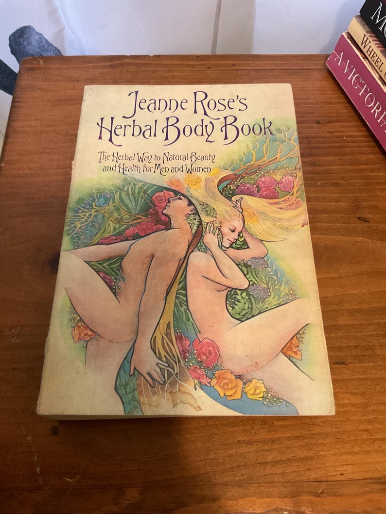 May include: A paperback book titled Jeanne Rose's Herbal Body Book: The Herbal Way to Natural Beauty and Health for Men and Women. The cover features a colorful illustration of two figures surrounded by green foliage, yellow hair, and flowers.