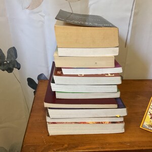 May include: A vertical stack of ten paperback books with varying spine colors including black, maroon, and white, positioned next to a rectangular yellow bookmark featuring the text "Fall Over You Fall In" printed in black and red lettering.
