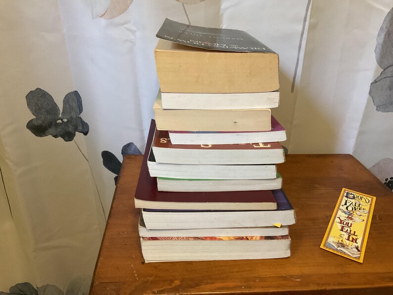 May include: A vertical stack of ten paperback books with varying spine colors including black, maroon, and white, positioned next to a rectangular yellow bookmark featuring the text "Fall Over You Fall In" printed in black and red lettering.