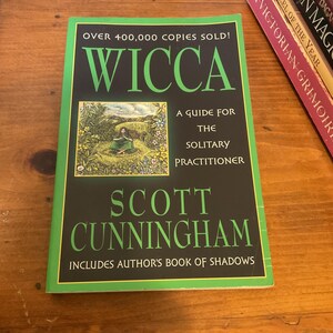 May include: A paperback book titled Wicca: A Guide for the Solitary Practitioner by Scott Cunningham. The black cover features green text, a small illustration of a person in a field, and the text: Over 400,000 copies sold! Includes author's Book of Shadows.
