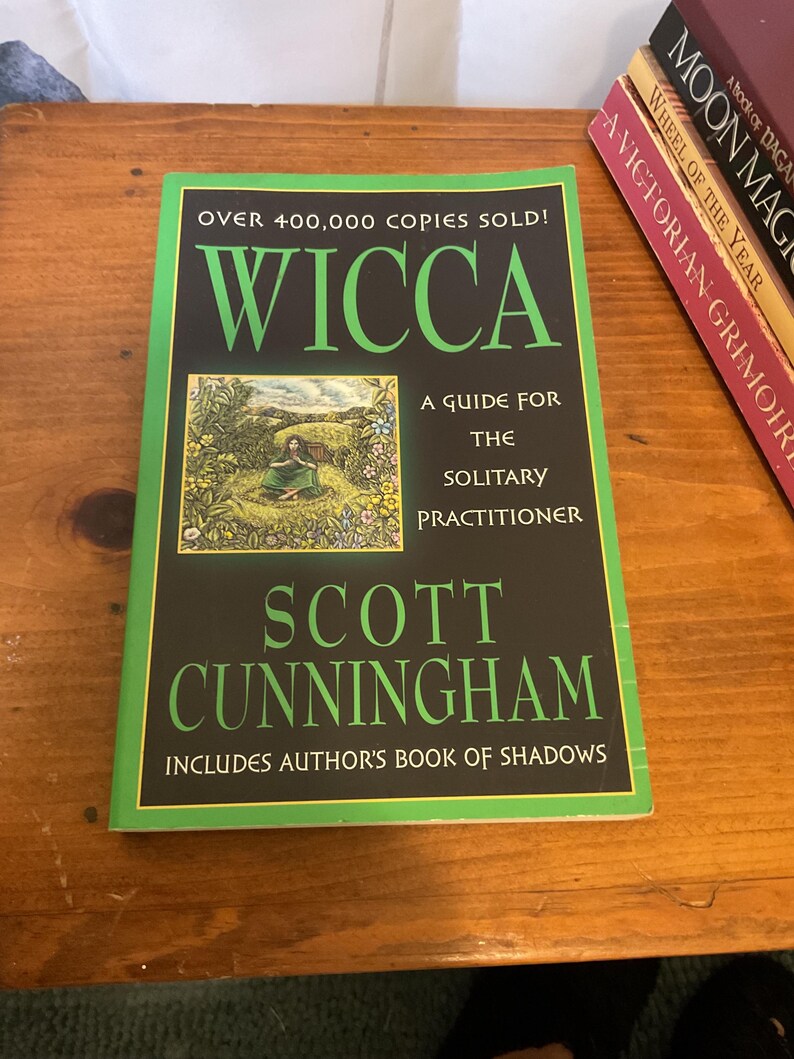 May include: A paperback book titled Wicca: A Guide for the Solitary Practitioner by Scott Cunningham. The black cover features green text, a small illustration of a person in a field, and the text: Over 400,000 copies sold! Includes author's Book of Shadows.
