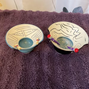 May include: Two decorative ceramic bowls with a white exterior and blue interior. Each bowl has a unique design with black lines and a small, round opening. One bowl has small figures attached. The other has two purple pencils with red tips.