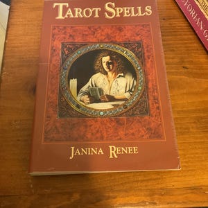 May include: A book titled Tarot Spells by Janina Renee with a dark red cover. The front features a circular illustration of a person holding tarot cards next to a lit candle, framed by a decorative border with small colored stones.