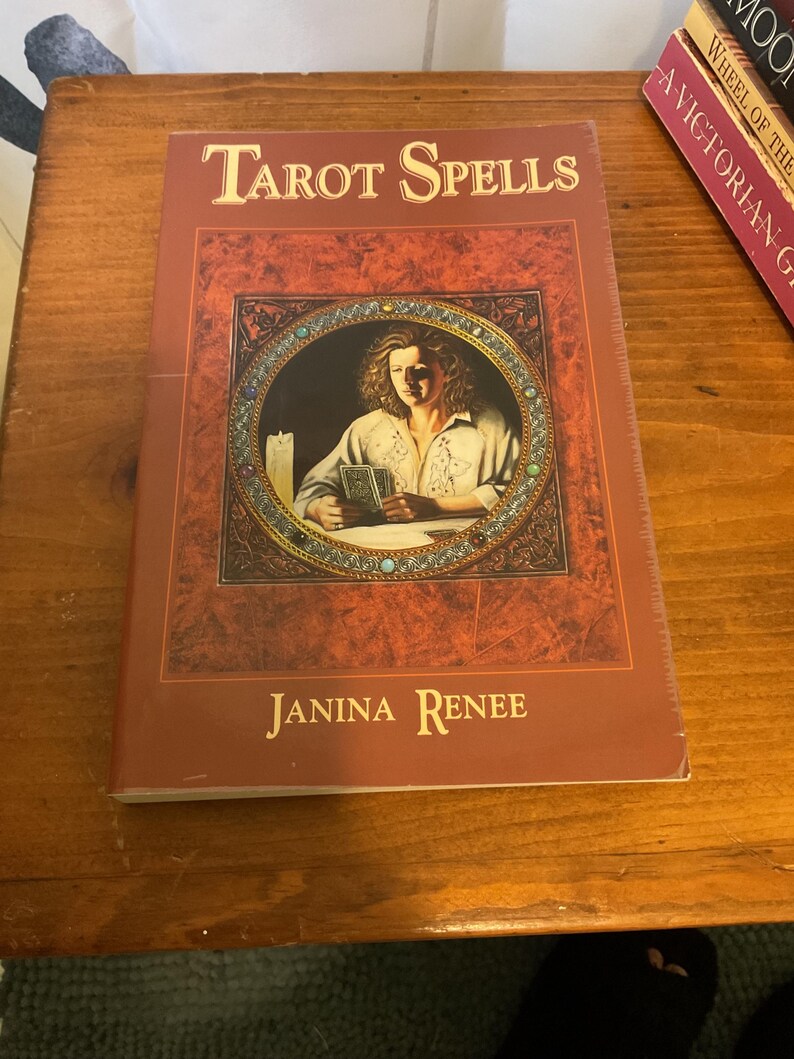 May include: A book titled Tarot Spells by Janina Renee with a dark red cover. The front features a circular illustration of a person holding tarot cards next to a lit candle, framed by a decorative border with small colored stones.