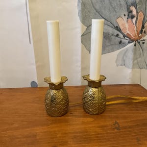May include: Two decorative pineapple-shaped candle holders with white candle inserts. The candle holders are gold-colored and sit on a wooden surface. The background includes a floral patterned curtain.