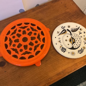 May include: Two decorative trivets. One is bright orange with a circular design of cut-out shapes. The other is white with a black and gold apple illustration, surrounded by floral and geometric patterns. Both are round.