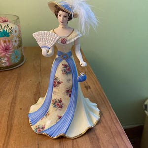 May include: A porcelain figurine of a woman in a Victorian-era gown. The dress is cream-colored with a floral pattern and blue accents. She wears a hat with feathers and holds a fan and a small purse.