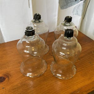 May include: Four clear glass candle holders with black tops are arranged on a wooden surface. The candle holders have a flared base and a rounded top, with a decorative edge. The background includes a white curtain and a floral patterned curtain.