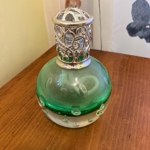 May include: A decorative glass bottle with a round, green body and a clear base. The bottle has a silver filigree top. The green section has air bubbles. The bottle sits on a wooden surface.