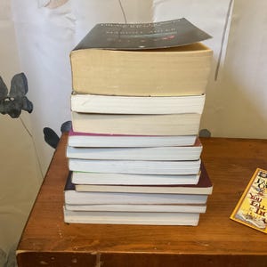 May include: A vertical stack of nine books with varying spine colors and page edges, topped with a black-covered book. Beside the stack lies a rectangular yellow bookmark featuring the text "Does it fall over? You fall in" in black lettering.