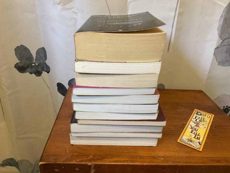 May include: A vertical stack of nine books with varying spine colors and page edges, topped with a black-covered book. Beside the stack lies a rectangular yellow bookmark featuring the text "Does it fall over? You fall in" in black lettering.