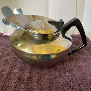 May include: A shiny, silver-colored teapot with a black handle and lid. The teapot has a rounded body and a spout. The handle is curved and attached to the side of the pot. The teapot is sitting on a purple towel.