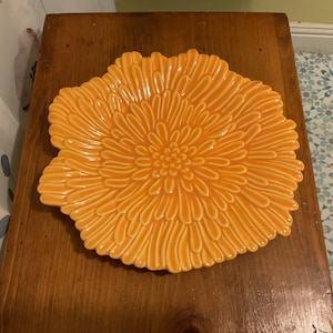 May include: An orange ceramic decorative plate in the shape of a flower. The plate has a detailed, textured design with petal-like ridges radiating from the center. The plate is on a wooden surface.