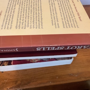 May include: A stack of three books. The top book is titled Tarot Spells by Janina Renee, featuring a cream-colored back cover with black text and a maroon spine. Below it is a thin maroon book, and the bottom book has a white spine and cover.